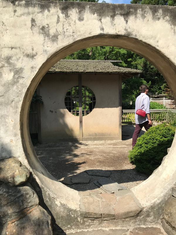 A large circle door in a stone wall opens onto another wall which has a small circle window. A tall man in a light shirt walks out of the scene to the right.