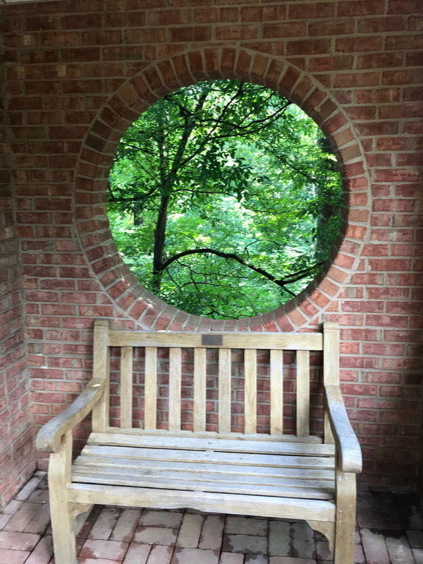 A circular window in a brick wall looks into a forest. A bench sits under the window.