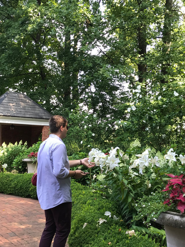 A tall man in a light colored shirt has his hand under a stalk of white lilies.